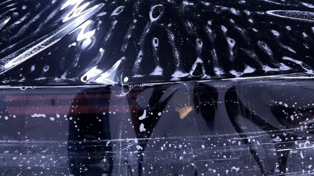 Employees of a car detailing studio apply clear protective PPF film on the hood of a black car. The video showcases the meticulous process ensuring high-quality protection and finish.