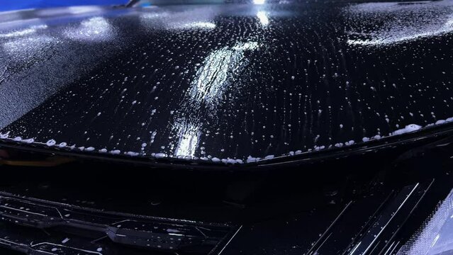 Employees of a car detailing studio apply clear protective PPF film on the hood of a black car. The video showcases the meticulous process ensuring high-quality protection and finish.