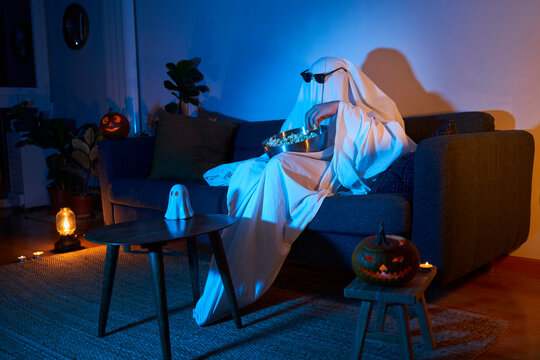Halloween scary movie night. Person in ghost costume with sunglasses and pumpkin decor eating popcorn while watching tv in sofa