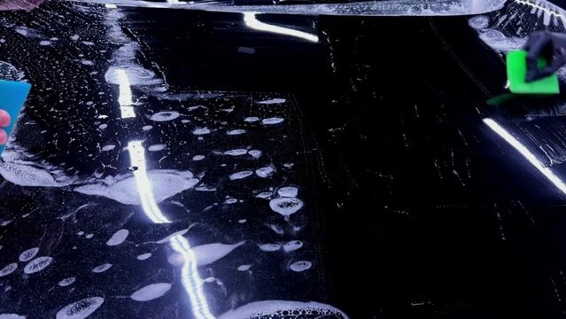 Employees of a car detailing studio apply clear protective PPF film on the hood of a black car. The video showcases the meticulous process ensuring high-quality protection and finish.