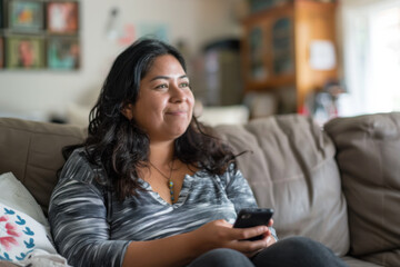 an image of an adult hispanic woman sits on a couch in her home, smiling and holding a smartphone. She is looking away, thinking about something
