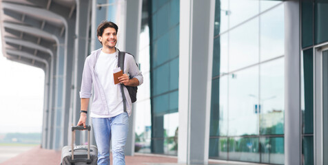 Traveling concept. Happy man in casual clothes walking outdoors near airport terminal with luggage, passport and tickets, ready for trip, free space