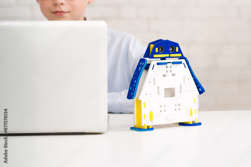 Aufkleber Boy is working on a laptop computer with a toy robot sitting on the desk beside them. The robot is made of white plastic and has blue and yellow accents.