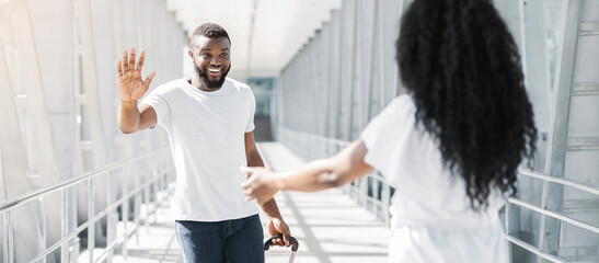 A smiling black man with a beard is standing in an airport terminal, waving hello to a woman walking towards him. He is holding a suitcase and looks happy to see her.