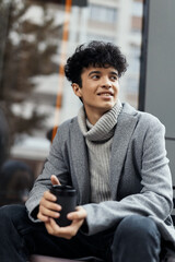 Fashionable young man smiling with a cup of coffee, sitting on a bench and looking at the camera