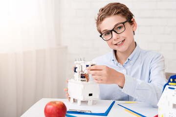 A young boy is sitting at a desk in his home, wearing glasses and a white shirt. He is smiling as he carefully assembles a white robot, focusing on the details of the construction.