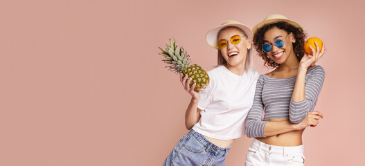 This photo shows two happy young women holding fruit while wearing hats and sunglasses. They are standing on a pink background and are smiling at the camera.