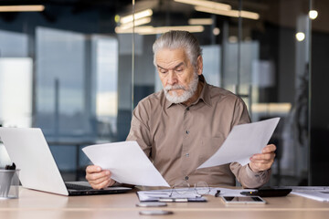 Older male professional analyzing paperwork while working on a laptop in a contemporary office...