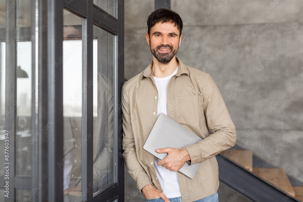 Poster a man in a casual shirt stands in a modern office setting, holding a laptop computer close to his bo
