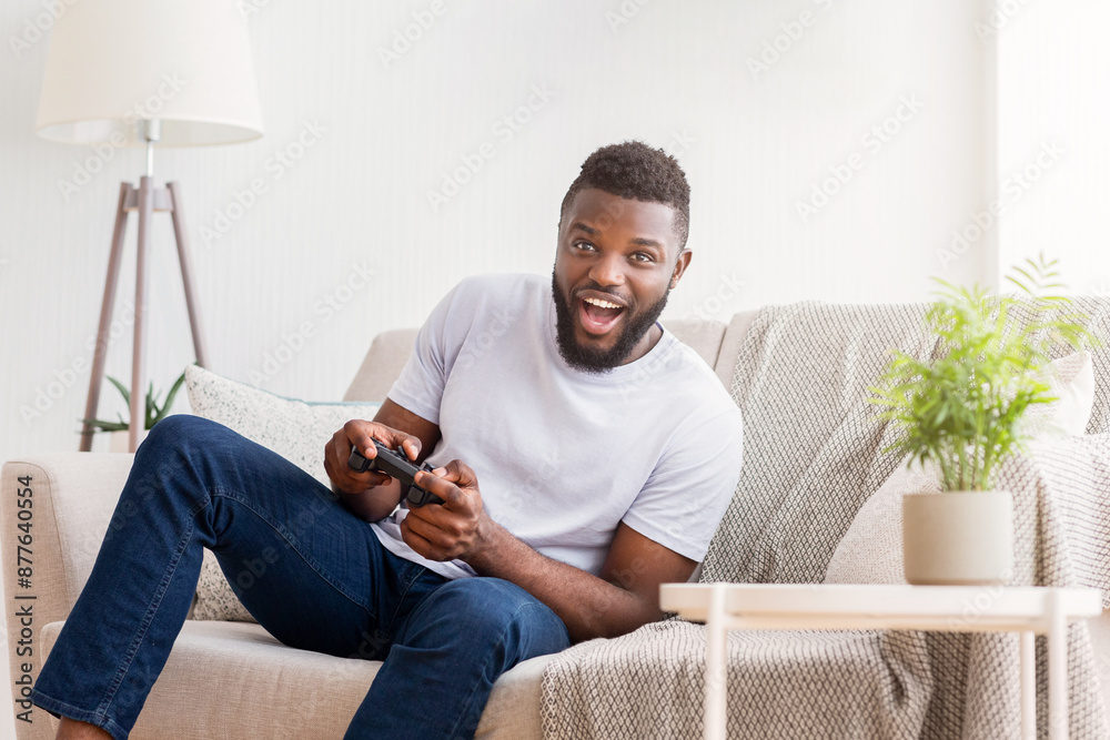 Wall mural a young black man sits on a couch in his living room, excitedly playing video games using a black co