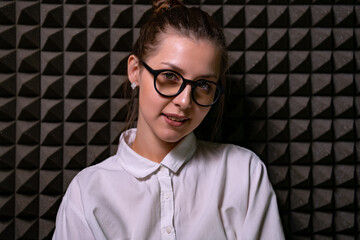 Close up of a happy young woman wearing glasses showing a smile to the camera against a dark wall.