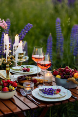 Beautiful elegant romantic dinner table decor, surprise day arrangement. Marriage proposal, engagement private party for two in a meadow. Sunset, golden hour. Cheese board, rose wine, fruits, lupine