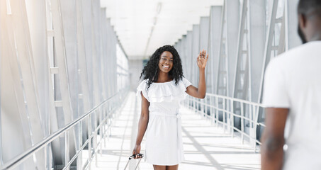 African American woman in a white dress walks down a terminal corridor at an airport, waving goodbye to someone out of frame. She is smiling and appears to be happy to be leaving.