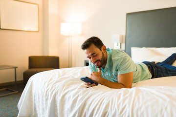 Happy young Caucasian man enjoying his smartphone while relaxing on a hotel bed