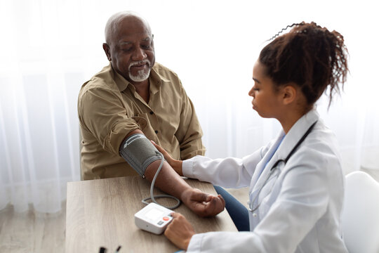 Hypertension In Older Age. Young Female Medical Worker Measuring Arterial Blood Pressure Of Senior Black Man Using Cuff, Patient Having Problems With Tension, Sitting At Table. Health Care Concept