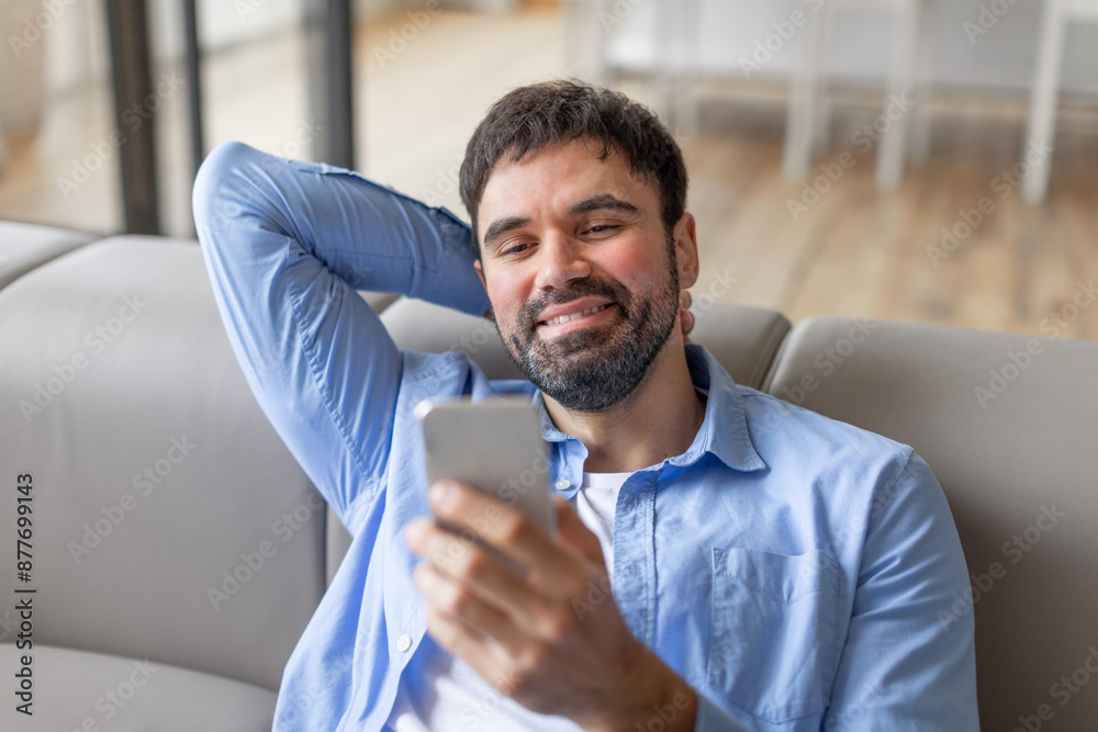 Poster smiling millennial bearded man reclining on couch at home, using smartphone, scrolling or chatting o