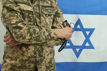 Fototapeta premium Man in military uniform with crossed arms and stethoscope against flag of Israel, closeup. Health care concept