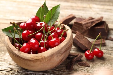 Fresh cherries with green leaves in bowl and dark chocolate on wooden table, closeup