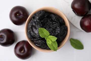 Delicious prunes with mint in bowl and fresh ripe plums on white table, flat lay