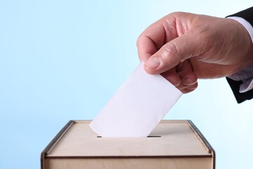 Man putting his vote into ballot box against light blue background, closeup