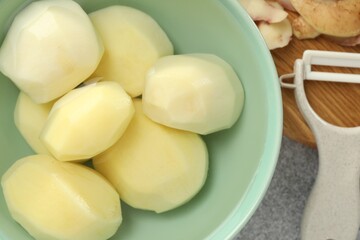 Fresh raw potatoes in bowl, peeler and peels on light grey table, top view