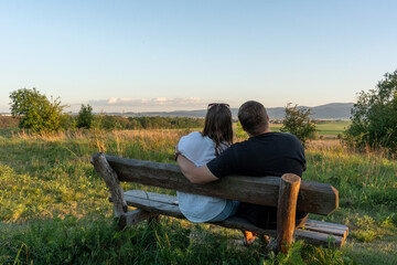 Couple sitting on wooden bench in field looking at sunset view on peaceful evening. Concept of...