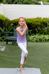 Fototapeta premium Vertical of happy biracial girl standing on one leg in garden, practicing yoga, with copy space
