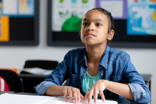 Blind biracial schoolgirl sitting at desk in class reading braille, with copy space - Powered by Adobe