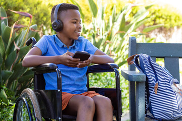 Obraz premium Joyful African American boy in wheelchair with headphones, smartphone in garden