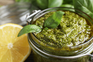 Tasty pesto sauce in glass jar, basil and lemon on table, closeup