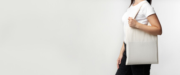 A woman in a white shirt and black pants holds a white tote bag against a white background.