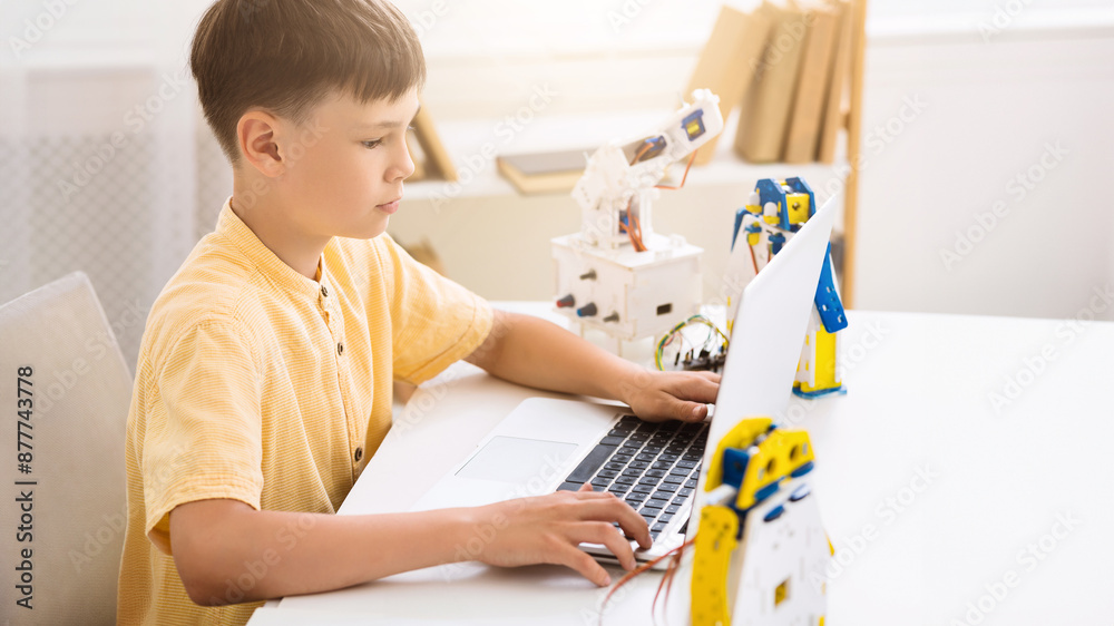 Aufkleber A young boy is learning about robotics by using a laptop computer. The boy is sitting at a table and is looking at the screen of the laptop. There is a robotic arm behind the laptop.
