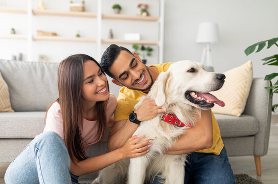 Portrait of happy multiracial couple scratching their pet dog, sitting on floor at home. Arab guy and his Caucasian girlfriend hugging their golden retriever in living room - Powered by Adobe