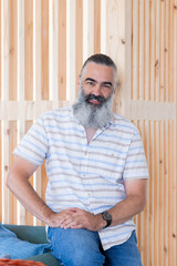 Smiling senior man with beard sitting on couch, wearing casual striped shirt and jeans