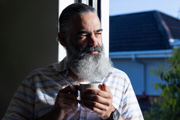 Holding cup, senior man with beard enjoying morning sunlight indoors