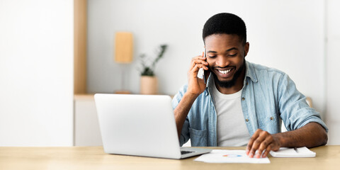 Fototapeta premium Cheerful african businessman checking financial reports and talking on phone, happy about results of his company. Copy space