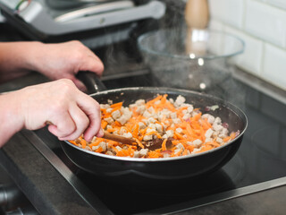The hand of a senior woman holds a wooden spoon and stirs the meat with carrots, frying in a pan