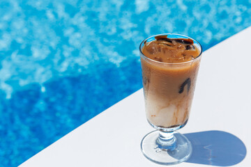 Iced coffee Frappe Fredo against blue clear water of the swimming pool, on white table . Summer minimalistic background, holiday or vacation concept. Sun and shadows. Copy space