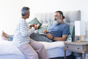 Obraz premium Senior woman giving medication to man using laptop while resting in bed