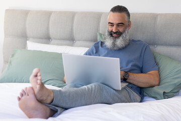 Working from home, senior man using laptop while sitting on bed