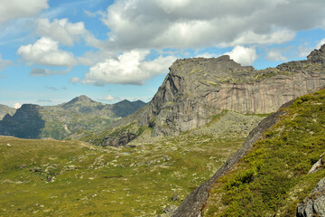 A fragment of a high rock in the shape of a man lying on his back surrounded by mountain ranges on a cloudy summer day.