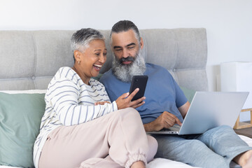 Using smartphone and laptop, senior couple relaxing on bed and smiling