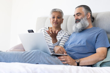 Using laptop and smartphone, senior couple relaxing on bed and discussing something