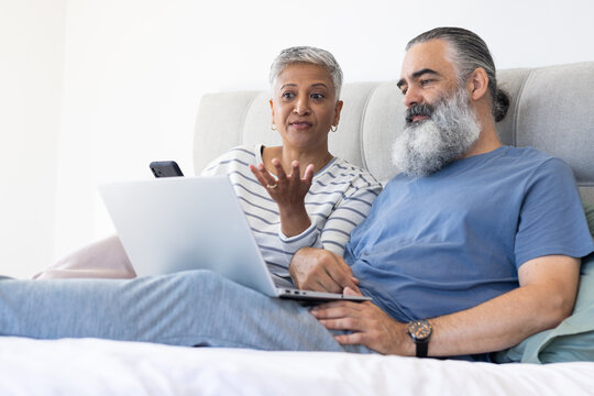 Using laptop and smartphone, senior couple relaxing on bed and discussing something