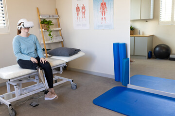 Woman in prosthetic leg using VR headset while sitting on medical examination table, copy space