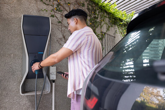Charging electric car, man using smartphone while connecting vehicle to charger