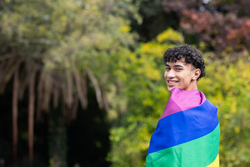 Smiling young man wrapped in rainbow flag, celebrating LGBTQ+ pride outdoors, copy space