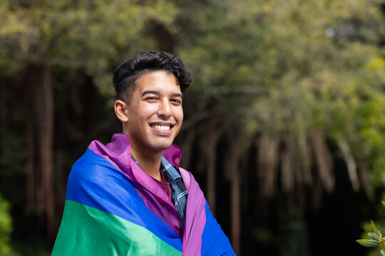Smiling man wrapped in pride flag, standing outdoors in nature, copy space