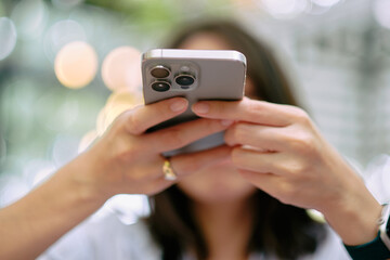 A woman is taking a photo of a slice of cake and a cold drink with her smartphone. She is sitting at a table outside, and the background is blurred. The focus is on the phone and the food.