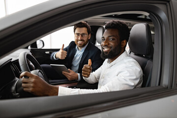 Happy customer and dealer showing thumbs up, expressing satisfaction and successful deal inside car dealership. Caucasian car dealer sit inside vehicle with African American buyer.